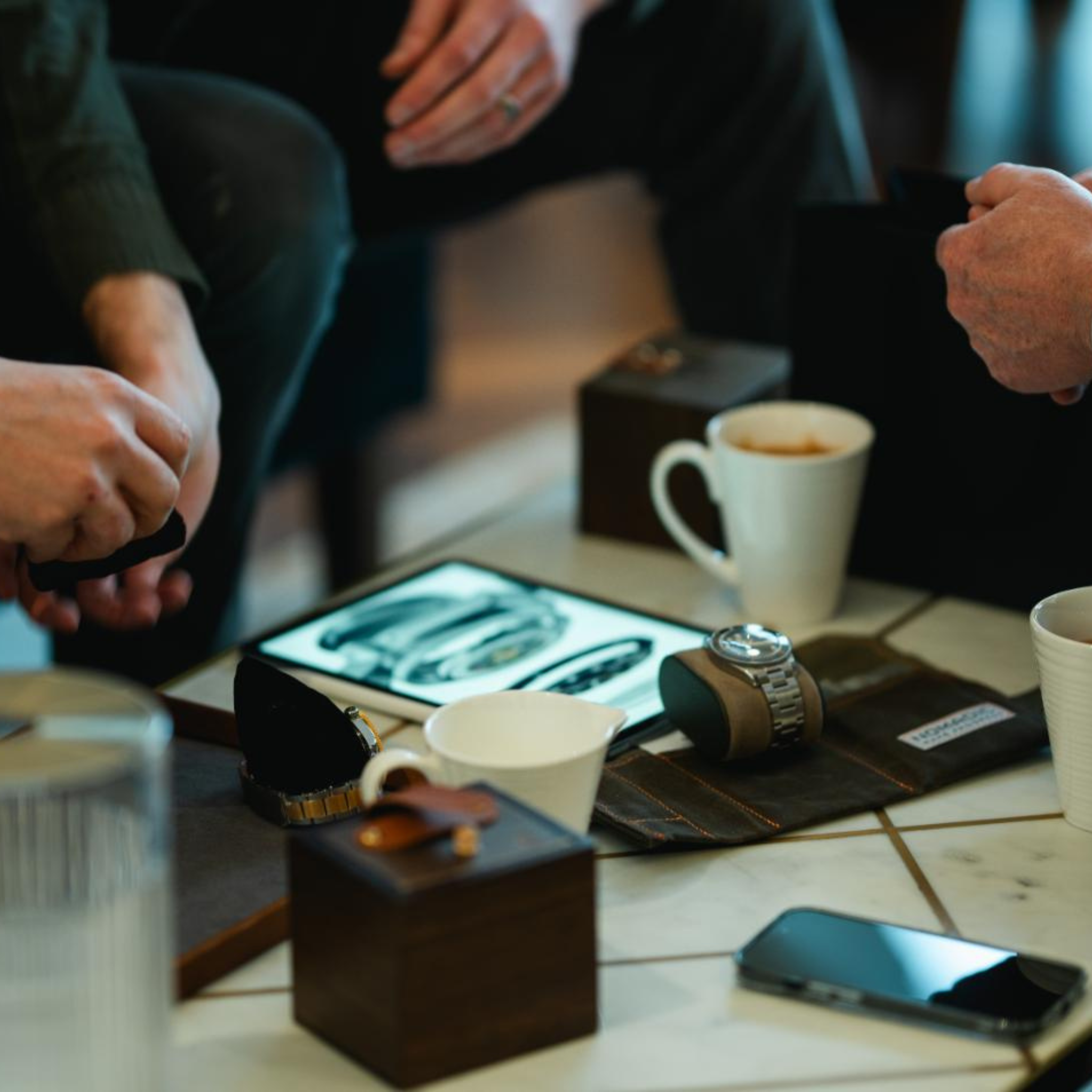 People examining wristwatches on a table with coffee cups and an open tablet displaying watch images.